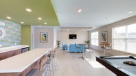 The Flats At 345 community lounge is a thoughtfully designed, single-floor amenity space that blends social functionality with contemporary style. The open-concept layout flows seamlessly from one zone to the next, creating an inviting environment for residents to gather, relax, and entertain.

At the heart of the space is a striking kitchenette area, distinguished by its bold green accent ceiling and a colorful subway tile backsplash in shades of blue, green, and gray. A large white island with counter seating anchors the space, while rich wood cabinetry, a stainless steel refrigerator, and an integrated sink provide full utility. The herringbone-patterned light wood flooring adds a refined touch throughout.

The lounge area features plush blue upholstered seating arranged around a round coffee table, complemented by a wall-mounted flat-screen television — ideal for casual gatherings. Framed colorful artwork adorns the walls, adding personality and warmth.

A games and casual dining zone rounds out the space, equipped with a pool table, high-top bistro tables with bar-height stools, and an additional wall-mounted television. Large windows with white blinds flood the area with natural light, creating a bright and energetic atmosphere. The overall design balances modern aesthetics with practical, community-centered amenities.
