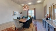 This single-story dining space makes a strong design statement through a carefully curated blend of contemporary art and eclectic furnishings. The room's elevated ceilings create an airy, open atmosphere, anchored by a dramatic sculptural copper chandelier that serves as the undisputed focal point. Light wood-look flooring runs throughout, providing a clean, neutral foundation that complements the space's bold decorative choices.

At the center of the room sits a substantial solid wood dining table surrounded by an intentionally mismatched yet cohesive arrangement of seating, including dark slipcovered chairs with fringe detailing, a patterned accent chair, and teal upholstered seats with decorative trim. A richly colored vintage-style area rug grounds the dining arrangement, adding warmth and texture.

Natural light enters through a large window dressed with two-toned curtains in rust and teal, enhancing the room's vibrant color palette. One wall features a built-in granite-topped sideboard flanked by large-scale abstract artwork in warm terracotta and teal tones, while additional abstract framed pieces adorn an adjacent wall. Glass-panel French doors connect the dining area to an adjoining space, reinforcing the open, flowing layout of this thoughtfully designed interior.