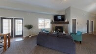 Located at 208 Catalpa Dr in Bloomington, Illinois, this single-story community clubhouse offers a well-appointed and versatile gathering space designed for comfort and functionality. The main lounge area is anchored by a tiled corner fireplace with a rich wood mantel and wall-mounted television, creating a warm and inviting focal point. Plush seating, including a large sectional sofa and accent chairs in teal, complements the neutral gray walls and patterned carpet flooring throughout. An arched-panel entry door adds a refined architectural touch to the entryway. Adjacent to the lounge, a casual dining area features high-top tables and bar-height seating, bathed in natural light from large windows that overlook an outdoor playground. A separate gathering room with vaulted ceilings and a bold dark accent wall is furnished with long communal tables and industrial-style stools, ideal for group activities or meetings. A ceiling fan adds to the airy feel of this room. The fully equipped kitchen includes warm wood cabinetry, granite-style countertops, stainless steel appliances, a double sink, and a long serving counter with bar seating — well-suited for catered events or communal meals. The space overall exudes a modern yet welcoming ambiance throughout.