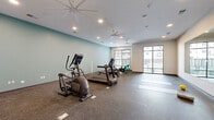 Located at 501 4th Ave SE in Cedar Rapids, Iowa, this fitness center offers a well-appointed, single-floor workout space designed to support a broad range of exercise routines. The room features durable, shock-absorbing rubber flooring throughout, making it suitable for both cardio and stretching activities. Recessed lighting and multiple large ceiling fans provide consistent illumination and airflow across the space.

The cardio equipment section includes a treadmill, elliptical trainer, rowing machine, and cycling bikes, providing users with a comprehensive selection of aerobic workout options. A dedicated yoga and stretching area is defined by exercise mats laid out on the open floor. A large wall mirror with an adjacent flat-screen television enhances the workout experience and allows users to monitor their form.

Natural light is a standout feature of this fitness center, with floor-to-ceiling windows fitted with adjustable blinds along multiple walls, offering views of the surrounding exterior. The walls are finished in a calming two-tone palette of soft sage green and neutral white, contributing to an energizing yet focused atmosphere. The overall design is clean, modern, and functional, making this fitness center a practical and inviting amenity for those seeking an active lifestyle in the heart of Cedar Rapids.