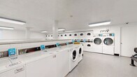 This single-floor communal laundry facility is designed to efficiently serve a high volume of users. The main laundry room is spacious and well-lit, featuring overhead fluorescent lighting that illuminates rows of front-loading and top-loading washing machines alongside stacked and standalone dryers. The machines are arranged in organized rows along the walls and in central islands, maximizing capacity throughout the room. Each unit is equipped with card or coin-operated payment panels, providing convenient and cashless operation. The floors throughout are finished with durable speckled vinyl, easy to maintain and well-suited for a high-traffic environment. The white walls and white cabinetry create a clean, functional aesthetic. A dedicated folding area provides counter space for sorting and folding laundry, accompanied by a waiting area furnished with sturdy black metal benches, offering a comfortable spot to rest during the laundry cycle. A secondary, smaller laundry room is also present, featuring additional washers positioned beneath a window that allows natural light to filter in. An exterior door provides direct outdoor access, and a fire extinguisher is visibly mounted for safety. Overall, the facility is designed with practicality and efficiency in mind.
