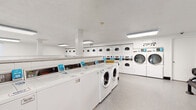 This single-floor communal laundry facility is designed to efficiently serve a high volume of users. The main laundry room is spacious and well-lit, featuring overhead fluorescent lighting that illuminates rows of front-loading and top-loading washing machines alongside stacked and standalone dryers. The machines are arranged in organized rows along the walls and in central islands, maximizing capacity throughout the room. Each unit is equipped with card or coin-operated payment panels, providing convenient and cashless operation. The floors throughout are finished with durable speckled vinyl, easy to maintain and well-suited for a high-traffic environment. The white walls and white cabinetry create a clean, functional aesthetic. A dedicated folding area provides counter space for sorting and folding laundry, accompanied by a waiting area furnished with sturdy black metal benches, offering a comfortable spot to rest during the laundry cycle. A secondary, smaller laundry room is also present, featuring additional washers positioned beneath a window that allows natural light to filter in. An exterior door provides direct outdoor access, and a fire extinguisher is visibly mounted for safety. Overall, the facility is designed with practicality and efficiency in mind.