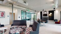 Located at 2981 Olentangy River Road in Columbus, Ohio, this thoughtfully designed amenity space offers a versatile, open-plan layout across a single floor. The main lounge area features plush seating arrangements, a large conference table, and stylish pendant lighting, creating a welcoming environment suited for both relaxation and collaboration. Exposed ductwork along the ceiling adds an industrial touch, while recessed lighting ensures the space remains bright and functional throughout.

A dedicated business center, finished in a vibrant teal, provides multiple computer workstations along wraparound white desks, complemented by built-in cabinetry and a multifunction printer — ideal for productivity needs. An adjoining room, also finished in teal, features additional workstations and a high-top table set against a large floor-to-ceiling window that frames lush greenery and fills the room with natural light.

The main open area connects to glassed-in conference and meeting rooms with dark-framed windows, offering semi-private spaces for group gatherings or focused work. Colorful wall art and decorative panels add personality throughout, while neutral carpet flooring ties the varied zones together cohesively. The overall design balances modern aesthetics with practical functionality, making this a dynamic and well-appointed communal space.