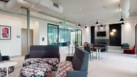 Located at 2981 Olentangy River Road in Columbus, Ohio, this thoughtfully designed amenity space offers a versatile, open-plan layout across a single floor. The main lounge area features plush seating arrangements, a large conference table, and stylish pendant lighting, creating a welcoming environment suited for both relaxation and collaboration. Exposed ductwork along the ceiling adds an industrial touch, while recessed lighting ensures the space remains bright and functional throughout.

A dedicated business center, finished in a vibrant teal, provides multiple computer workstations along wraparound white desks, complemented by built-in cabinetry and a multifunction printer — ideal for productivity needs. An adjoining room, also finished in teal, features additional workstations and a high-top table set against a large floor-to-ceiling window that frames lush greenery and fills the room with natural light.

The main open area connects to glassed-in conference and meeting rooms with dark-framed windows, offering semi-private spaces for group gatherings or focused work. Colorful wall art and decorative panels add personality throughout, while neutral carpet flooring ties the varied zones together cohesively. The overall design balances modern aesthetics with practical functionality, making this a dynamic and well-appointed communal space.