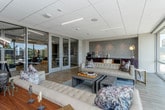 This single-floor commercial space combines contemporary design with functional versatility, offering a polished environment suited for professional or collaborative use. The layout is divided into distinct yet interconnected zones, unified by light wood flooring and a neutral, modern color palette accented by deep navy blue walls.

The lounge area serves as a central gathering space, furnished with tufted leather sofas, a layered area rug in cool blue tones, and a rustic wood coffee table. Floor-to-ceiling glass windows flood the area with natural light while providing broad views of the surrounding streetscape and urban environment. A built-in beverage station with dark cabinetry and a textured tile backsplash is conveniently positioned adjacent to the lounge.

The conference and work area features a long white communal table with modern seating, pendant lighting, and wall-mounted displays. Glass partition walls with aluminum framing connect this zone to the lounge, maintaining visual openness throughout the space.

Recessed and linear LED lighting complements the drop-tile ceiling, ensuring consistent illumination across all areas. The overall ambiance is clean, bright, and professional, with a design that balances comfort and productivity.