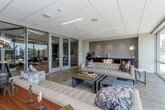 This single-floor commercial space combines contemporary design with functional versatility, offering a polished environment suited for professional or collaborative use. The layout is divided into distinct yet interconnected zones, unified by light wood flooring and a neutral, modern color palette accented by deep navy blue walls.

The lounge area serves as a central gathering space, furnished with tufted leather sofas, a layered area rug in cool blue tones, and a rustic wood coffee table. Floor-to-ceiling glass windows flood the area with natural light while providing broad views of the surrounding streetscape and urban environment. A built-in beverage station with dark cabinetry and a textured tile backsplash is conveniently positioned adjacent to the lounge.

The conference and work area features a long white communal table with modern seating, pendant lighting, and wall-mounted displays. Glass partition walls with aluminum framing connect this zone to the lounge, maintaining visual openness throughout the space.

Recessed and linear LED lighting complements the drop-tile ceiling, ensuring consistent illumination across all areas. The overall ambiance is clean, bright, and professional, with a design that balances comfort and productivity.