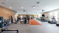 Located at 16152 SE 77th Court Rd in Summerfield, Florida, this single-story fitness center offers a comprehensive workout facility thoughtfully divided into two distinct exercise zones. The main workout area is a spacious, open-plan room featuring durable speckled carpet flooring, multiple ceiling fans, and recessed lighting throughout. A standout feature is the bold orange turf track running down the center of the floor, ideal for functional training and agility drills. This primary zone is outfitted with an extensive array of strength equipment, including cable machines, multi-station cable racks, weight benches, power racks, and a full dumbbell rack. A row of True-brand treadmills and cardio machines lines one wall, accompanied by wall-mounted flat-screen TVs. Large mirrors reflect the space, enhancing the sense of openness. An inspirational Muhammad Ali quote is painted prominently on a warm amber-toned accent wall, adding motivational character to the environment. Natural light filters in through multiple windows. The secondary room features light hardwood flooring, a ceiling fan, large mirrors, and abundant natural light from oversized windows. This area is dedicated to group fitness and functional training, housing spin bikes, TRX suspension systems, kettlebells, medicine balls, and resistance accessories, creating an inviting and versatile studio-style environment.