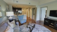 This single-floor residence showcases an open-concept design that seamlessly connects the living area and kitchen. The living space is anchored by a plush, light-toned area rug and features a comfortable sofa paired with a distinctive blue patterned accent chair, complemented by a sculptural glass-top coffee table that adds a refined focal point to the room.

The kitchen is appointed with dark cabinetry contrasting against light countertops, stainless steel appliances including a French door refrigerator, and pendant lighting that hangs above the counter bar seating area. Recessed lighting throughout provides consistent illumination across the open floor plan.

Rich dark hardwood flooring flows through the main living and kitchen areas, adding warmth and visual continuity. Multiple doorways are visible from the main space, suggesting well-organized secondary rooms. One doorway reveals what appears to be a laundry area with stacked or side-by-side units behind louvered doors. The overall ambiance is contemporary and cohesive, with a neutral color palette punctuated by carefully chosen furnishings and fixtures.