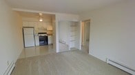 This single-floor unit offers a straightforward, functional layout designed to maximize usability within a compact footprint. The living area is finished with wall-to-wall neutral carpeting, creating a warm and comfortable atmosphere. A standout feature is the built-in storage closet with mirrored sliding doors and open shelving, providing both practical storage and a sense of added depth to the space.

Adjacent to the living area, the kitchen is efficiently arranged and appointed with white cabinetry, a white refrigerator, an electric range with an oven, and a dishwasher. The light-colored countertops and white finishes give the kitchen a clean, bright appearance. A ceiling fan with a light fixture overhead provides both ventilation and ambient lighting to the kitchen area.

A hallway visible from the main living space leads to additional rooms, suggesting a thoughtful separation of living and private areas. The overall color palette throughout is light and neutral, with white walls and trim contributing to an airy feel. The space is well-suited for those seeking a clean, low-maintenance layout with practical storage and essential amenities all contained on a single level.