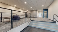 Located at 955 W Clairemont Ave in Eau Claire, Wisconsin, this expansive single-floor hallway corridor offers a thoughtfully designed commercial space that balances functionality with modern aesthetics. The corridor stretches generously, spanning a substantial footprint that allows for both circulation and informal gathering.

The hallway features a neutral color palette of warm beige and gray tones, accented by bold navy blue architectural details that add visual interest throughout. Underfoot, a patterned commercial carpet in muted blue-gray tones provides a durable yet attractive finish. The suspended tile ceiling houses recessed lighting that maintains consistent, even illumination throughout the length of the corridor.

Floor-to-ceiling windows line one side of the hallway, flooding the interior with natural light and offering open views of the surrounding green lawn and adjacent buildings. These large-format windows create a bright, welcoming atmosphere throughout the day.

Along the corridor, multiple informal seating areas are integrated seamlessly into the flow of the space, including pod-style seating units, lounge chairs, and small tables with chairs — ideal for collaboration or individual use. A split-level entry area features sleek metal cable railings alongside wide steps, adding architectural dimension to the space.

Glass-partitioned rooms accessible from the corridor provide transparency while maintaining distinct functional zones. The overall design reflects a clean, contemporary commercial environment well-suited for institutional or professional use.