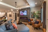 This single-floor community space is designed to impress, offering a thoughtfully curated open-concept layout that seamlessly blends multiple functional zones into one cohesive, modern environment. The grand entrance lobby sets a welcoming tone with soaring ceilings, exposed ductwork, striking round chandelier lighting, and distinctive stone columns that frame the space with architectural character. Light hardwood flooring flows consistently throughout, tying each area together with warmth and continuity.

The lounge area serves as the social heart of the space, featuring plush sectional sofas, eclectic throw pillows, a cowhide area rug, and rustic wooden accent tables. A large wall-mounted television anchors the room above a sleek linear fireplace set into a textured stone feature wall, while a tray ceiling with an artistic pendant chandelier adds architectural depth overhead. French doors open to the outdoors, allowing natural light to pour in.

Adjacent to the lounge, a dedicated coffee bar area is outfitted with slate-toned cabinetry, white subway tile backsplash, stainless steel appliances, and a bar-height island with stylish pendant lighting. A boldly colored accent wall designates the coffee station as its own distinct destination. Further into the space, a game zone with forage table, a dedicated workstation area, and additional lounge seating complete this versatile, amenity-rich environment.