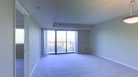 This single-floor space features an open living area finished with wall-to-wall carpeting in a neutral tone, contributing to a calm and understated ambiance. The room is defined by a set of floor-to-ceiling sliding glass doors that serve as the focal point of the space, flooding the interior with natural light and providing direct access to a private balcony enclosed by a metal railing. The outdoor views visible through the glass doors include surrounding greenery, adding a sense of openness to the interior. A stepped ceiling detail frames the area near the balcony, adding a subtle architectural element that breaks up the otherwise straightforward layout. Neutral-toned walls complement the light-colored carpet, creating a cohesive and clean aesthetic throughout. HVAC vents are positioned both on the ceiling and near the baseboard along the wall, ensuring climate control throughout the space. White baseboards run along the perimeter of the room, providing a clean finish. The overall ambiance is bright, open, and functional, with the large glass doors serving as the primary design feature connecting the interior to the outdoors.
