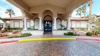 This single-story property presents a striking Mediterranean-inspired exterior, characterized by a covered portico supported by white columns, sweeping arched openings, and warm stucco façades in a sandy beige tone. A decorative stamped concrete driveway leads to the elegant double-door entrance, flanked by manicured tropical plantings, small palms, and low hedges that add curb appeal and a resort-like atmosphere.

Inside, the main common area showcases a contemporary interior design that contrasts thoughtfully with the traditional exterior. Teal accent walls and crisp white trim frame a spacious, open-plan layout with light wood-look flooring throughout. A central seating arrangement features tufted wingback chairs grouped around a round marble-top coffee table, anchored by a sleek, modern double-sided gas fireplace set into a dark feature wall. Above, a dramatic cascading chandelier draws the eye upward, emphasizing the generous ceiling height.

Adjacent to the lounge area, a dining space with multiple tables and chairs extends naturally from the main gathering zone. A "Wall of Honor" display adds a personalized, community-oriented touch. The overall ambiance is polished, welcoming, and well-appointed.