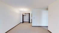 This single-floor space offers a clean and functional layout designed with simplicity and practicality in mind. The interior is characterized by crisp white walls and neutral-toned carpeted flooring, creating a bright and understated ambiance throughout.

The primary bedroom is a well-proportioned room featuring a dedicated closet with a built-in shelving system and a hanging rod, offering ample storage and organizational versatility. A pendant light fixture hangs above, providing focused overhead lighting that complements the room's understated aesthetic. Dark-framed door surrounds provide a subtle contrast against the white walls, adding a touch of visual definition to the space.

The room also features a partial wall partition that adds architectural interest while subtly defining areas within the open layout. Base molding in a dark finish runs along the perimeter of the room, reinforcing the clean, minimal design language present throughout the space. Electrical outlets are conveniently positioned along the walls to accommodate a variety of furniture arrangements. Overall, the space presents a calm, neutral palette that serves as a versatile backdrop, ready to be personalized to suit a range of interior styles and preferences.