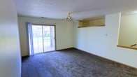 This single-floor space features a spacious living area finished with wall-to-wall carpeting in a neutral tone, creating a comfortable and cohesive feel underfoot. The room is illuminated by natural light streaming through a large sliding glass door, which provides outdoor access and views of a green exterior space beyond. A brass ceiling fan is centrally mounted on the textured popcorn ceiling, serving both a functional and decorative purpose while contributing to air circulation throughout the room. Along one wall, a built-in horizontal shelf adds a practical architectural detail, offering display or storage potential without occupying floor space. The walls are painted in a soft, light yellow tone, complemented by warm wood-toned baseboards that frame the room cleanly. The overall layout is open and unobstructed, allowing for flexible furniture arrangements to suit a variety of uses. The combination of natural light, neutral finishes, and thoughtful built-in features gives the space a straightforward, livable character suited to everyday comfort.
