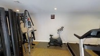 This fitness center is a dedicated single-room workout space designed to provide residents or guests with a convenient exercise option. The room features white walls and carpeted flooring, with a protective rubber mat placed beneath the equipment to preserve the floor surface. Recessed lighting in the ceiling provides adequate illumination throughout the space.

The fitness center is equipped with a multi-use cable and weight machine positioned along the left wall, offering a variety of strength training exercises. A Life Fitness upright stationary bike occupies the central area of the room, providing a solid option for low-impact cardiovascular workouts. A treadmill is also present along the right side, catering to those looking for walking or running exercises. A fitness center information sign is posted on the wall, providing guidelines for use of the facility.

The overall layout of the space is straightforward and functional, making efficient use of the available square footage. While compact, the room accommodates a well-rounded selection of equipment suited for both cardio and strength training, offering users a practical and accessible fitness facility.