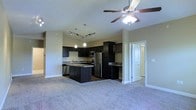 This single-floor residence showcases an open-concept layout that seamlessly connects the kitchen and living area, creating a cohesive and functional living environment. The kitchen is a standout feature, equipped with rich dark cabinetry that extends to the upper walls, offering ample storage throughout. Granite-style countertops provide durable work surfaces, while stainless steel appliances, including a refrigerator and range with overhead microwave, add a modern, polished touch. A central island enhances the kitchen's practicality, offering additional counter space and serving as a natural dividing point between the kitchen and the adjoining living area. Pendant lighting hangs above the island, complementing a track lighting system that illuminates the kitchen workspace. The living area features neutral-toned carpet underfoot and is equipped with a ceiling fan fitted with a light fixture, promoting air circulation and comfort. Soft, neutral wall tones throughout maintain a clean, understated aesthetic. A doorway visible from the main space hints at additional rooms beyond, suggesting a thoughtfully segmented floor plan that balances open living with private spaces.