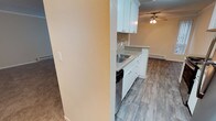 This single-floor space offers a well-organized layout that seamlessly connects the kitchen and living areas. The kitchen is appointed with crisp white cabinetry, light granite-style countertops, and a stainless steel sink, complemented by a classic subway tile backsplash that adds a timeless, clean aesthetic. A built-in dishwasher provides added convenience. The kitchen opens to the adjoining living area, which features wood-look plank flooring that lends warmth and durability to the space. A ceiling fan is centrally positioned in the living area, promoting air circulation and comfort year-round. Baseboard heating runs along the perimeter, ensuring efficient climate control throughout the colder months. The transition between the kitchen and living space is defined by a partial wall, creating a sense of separation while maintaining an open, connected feel. Neutral wall tones and a textured ceiling contribute to a calm, understated ambiance throughout. The combination of updated finishes, practical amenities, and a cohesive design makes this a highly functional and inviting space suited for a variety of living arrangements.