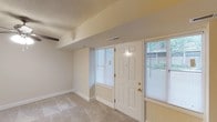This single-floor space presents a clean, functional layout designed for comfortable everyday living. The entry area sets a welcoming tone with a classic six-panel white front door flanked by large windows fitted with horizontal blinds, allowing abundant natural light to fill the interior while maintaining privacy. A sidelight window beside the entry door further enhances the brightness of the space and offers a view of the surrounding greenery outside.

The interior features neutral beige walls that create a calm, understated atmosphere, complemented by light-toned carpeted flooring that runs throughout the space. A textured popcorn ceiling adds a classic residential character overhead, while a low-profile soffit detail with a built-in HVAC vent provides practical climate control. The visible ceiling beam accent adds a subtle architectural element that defines the transition between areas.

The walls are clean and unadorned, providing a versatile canvas for personalization. Baseboards are neatly finished throughout, contributing to the well-maintained appearance of the space. Overall, this residence offers a straightforward, move-in-ready environment with ample natural light, modest architectural detailing, and a neutral palette that suits a wide range of interior styles.