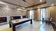 This single-floor game and lounge space is designed with both function and aesthetics in mind. The centerpiece is a full-size billiard table in a dark wood finish, positioned beneath a striking rectangular industrial-style pendant light fitted with exposed Edison bulbs, providing focused illumination over the playing surface. The ceiling features recessed lighting and decorative tray detailing with warm wood trim accents, adding architectural depth to the room.

One wall is adorned with a bold geometric textured panel in neutral tones, serving as a sophisticated backdrop to a built-in bench seating area with dark upholstery and decorative throw pillows in navy and patterned fabrics. This lounge nook offers comfortable spectator seating directly adjacent to the billiards table.

Floor-to-ceiling windows and sliding glass doors line the exterior wall, flooding the space with abundant natural light and offering views of the surrounding outdoor greenery. Sheer and linen-tone drapery panels frame the windows elegantly. The flooring features smooth polished concrete layered with a large area rug in muted earth tones, enhancing the room's modern yet inviting ambiance. Bar-height seating is also visible near the window area.