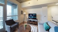 This single-floor space combines contemporary design with functional living across several well-appointed areas. The open-concept kitchen is outfitted with crisp white cabinetry, granite countertops, and stainless steel appliances, including a range, microwave, and sink — offering ample storage and prep space that flows seamlessly into the living area.

The living room features light hardwood-style flooring, a large window that draws in natural light, and an exposed concrete ceiling that lends an industrial character to the space. A media console, seating area, and floor lamp create a comfortable, casual atmosphere ideal for relaxing or entertaining.

The bedroom continues the warm, neutral palette with carpeted flooring and a distinctive exposed concrete ceiling detail overhead. The room is thoughtfully arranged with bedside tables and decorative wall art, fostering a calm, restful environment.

The bathroom is finished with large-format tile flooring in soft beige tones and features a glass-enclosed walk-in shower with white subway tile surround and chrome fixtures. A toilet and granite-topped vanity complete the space, maintaining the clean, modern aesthetic found throughout the unit.
