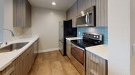 This single-floor residence offers a thoughtfully designed open-concept layout that seamlessly connects the living and kitchen areas. The kitchen is a standout feature, showcasing modern wood-grain cabinetry, stainless steel appliances including a refrigerator, electric range, and over-the-range microwave, complemented by a striking teal subway tile backsplash and crisp white countertops. A peninsula with a built-in sink extends into the living space, providing both additional prep area and a natural divide between the kitchen and the main living area. The living space is generously proportioned, featuring warm wood-look flooring and a distinctive white brick accent wall that adds texture and character to the room. Natural light filters in through shuttered windows, enhancing the bright, airy ambiance. The bathroom is clean and contemporary, featuring a tiled tub-and-shower combination, a floating wood-grain vanity with a white countertop, an undermount sink with brushed nickel fixtures, and a large mirror. A laundry area is also present within the unit. The overall design palette of neutral tones, modern finishes, and warm flooring creates a cohesive and inviting atmosphere throughout the space.