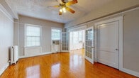 This generously sized 1-bedroom, 1-bathroom unit is spread across a single floor and showcases a thoughtful layout designed for comfortable living. The main living area is expansive and bathed in natural light from multiple windows, highlighting the warm, honey-toned hardwood floors that extend throughout the space. A ceiling fan provides both style and function overhead, while classic white trim and neutral gray walls create a clean, timeless aesthetic. Elegant French doors with glass pane detailing connect the living area to a bright, sun-filled secondary space, adding architectural character and an open, airy feel. The kitchen features white cabinetry, dark granite-style countertops, a double stainless steel sink, a gas range, and a striking mosaic tile backsplash that adds a decorative touch. A pass-through counter with seating connects the kitchen to the main living area, making it practical for everyday use. The bathroom retains a classic vintage charm, showcasing bold black-and-white diamond-patterned wall tiles, a full soaking bathtub with a shower, and a pedestal sink. A window in the bathroom allows for natural light and ventilation. Radiator heating units are present throughout, reflecting the unit's traditional character while ensuring year-round comfort.