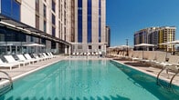 Located in the heart of downtown Phoenix, this rooftop pool deck offers a sophisticated urban retreat with sweeping views of the city skyline. The centerpiece is a generously sized, crystal-clear swimming pool flanked on all sides by rows of sleek white lounge chairs and umbrellas, ideal for sun-soaking beneath the Arizona sky. Elegant white pergolas provide partial shade and architectural definition to the deck, complementing the clean, modern aesthetic throughout.

The terrace features distinct zones designed for relaxation and socializing. Lush artificial turf areas are furnished with contemporary outdoor seating arrangements—including woven rattan chairs, plush sofas, and natural wood accent tables—offering comfortable, lounge-style gathering spaces. Frosted glass privacy screens partition select seating areas for a more intimate experience. Floor-to-ceiling glass walls border portions of the deck, reflecting the pool and skyline while seamlessly connecting interior spaces to the outdoor environment.

The overall design is crisp and contemporary, with large-format pavers, white and neutral tones, and a clean architectural language that emphasizes openness. Panoramic views of the surrounding downtown Phoenix high-rises and the broader urban landscape frame the space on all sides, creating a vibrant, resort-caliber rooftop experience in the city center.