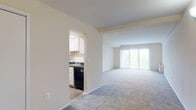 This single-floor unit offers a practical and functional layout with a neutral color palette throughout. The spacious living area is carpeted in a warm gray tone and is anchored by a large sliding glass door fitted with vertical blinds, allowing generous natural light and outdoor views to fill the room. The open pass-through connection to the kitchen creates a sense of flow between the two spaces.

The kitchen is efficiently designed with white cabinetry, dark granite-style countertops, a subway tile backsplash, and black appliances including a range, dishwasher, and full-size refrigerator. Wood-look flooring grounds the kitchen, providing a practical contrast to the carpeted living areas. A doorway from the kitchen leads back into the main living space, reinforcing the unit's open, connected feel.

The bedrooms are carpeted and finished in the same warm neutral tones, each featuring windows with blinds that provide natural light and privacy. Baseboard heating units are present in the bedrooms. The bathroom includes a full soaking tub with white tile surround and chrome fixtures, offering a clean and classic finish. Overall, the space presents a straightforward, move-in ready layout.