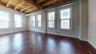 This single-floor loft showcases a striking industrial aesthetic defined by its exposed wooden beam ceilings with visible ductwork running throughout the open living areas. Rich, dark-stained hardwood floors extend across the main living and kitchen spaces, creating a warm contrast against the crisp white walls and structural wood columns that punctuate the interior.

The open-concept living area is generously proportioned, with multiple large windows fitted with horizontal blinds that invite abundant natural light and urban views. The kitchen is well-appointed with rich cherry wood cabinetry, granite-style countertops, and a full suite of stainless steel appliances including a refrigerator, electric range, and microwave. Pendant lighting above the kitchen counter adds a refined touch to the industrial setting.

The bedroom features plush carpeting, exposed beam ceilings, and a sliding-door closet with dual hanging rods and shelving for organized storage. A second carpeted room also benefits from tall windows with urban views. The bathroom includes a walk-in shower with a built-in bench and tile flooring. Overall, the space balances raw architectural character with functional, modern finishes.