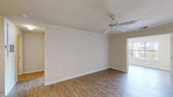 Located at 3808 Banyan Grove Ln in Virginia Beach, Virginia, this single-floor, one-bedroom unit offers a straightforward and functional layout suited for comfortable everyday living. The open living area is finished with wood-look flooring and features crown molding, a ceiling fan, and neutral-toned walls that create a bright, clean atmosphere. Natural light filters in through windows that offer views of the surrounding exterior, enhancing the overall sense of openness. A pass-through counter connects the living space to the kitchen, which is equipped with white cabinetry, a coil-burner electric range, dishwasher, and a full-size refrigerator, all set above matching wood-look flooring. The entryway features tile flooring and a solid paneled front door. The bedroom is carpeted, offering a softer underfoot feel, and includes a walk-in closet with built-in rod storage. Two windows in the bedroom provide ample natural light and outdoor views. The bathroom includes a single-sink vanity with a large mirror, a tiled soaking tub and shower combination, and consistent wood-look flooring. A door visible from the living area suggests access to a private outdoor space.