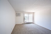 This single-floor residential unit offers a well-organized layout with distinct living, sleeping, and cooking areas. The living room is generously sized, finished with wall-to-wall carpet, a textured ceiling, and a large sliding glass door fitted with vertical blinds that floods the space with natural light and provides outdoor access. Adjacent to the living area, the galley-style kitchen is efficiently designed with warm oak cabinetry lining both walls, light-toned countertops, a double sink, tile flooring, and white appliances including a range and refrigerator. The bedrooms are carpeted and bright, each featuring ceiling fans with integrated lighting, windows with horizontal blinds that bring in natural views of surrounding greenery, and baseboard heating. One bedroom includes a built-in closet with sliding wood-panel doors and direct access to an adjoining room or bathroom. Throughout the unit, consistent warm wood trim accents the windows, doors, and baseboards, contributing to a cohesive and understated interior aesthetic. The textured ceilings are a recurring architectural detail across the living and sleeping spaces. Overall, the unit presents a functional and comfortable floor plan suited for everyday residential living.