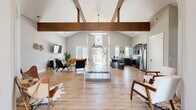 Located at 1125 Old Graves Mill Rd in Lynchburg, Virginia, this single-floor community space offers a generously sized, open-concept layout designed for both gathering and productivity. The centerpiece of the space is a dramatic vaulted ceiling accented by striking exposed wood beams, creating a warm yet contemporary architectural aesthetic throughout the main area. Light wood-tone flooring runs seamlessly underfoot, complementing the neutral gray walls and bright, airy atmosphere achieved through abundant natural light from multiple large windows and sliding glass doors.

The main open area flows effortlessly between a well-appointed lounge zone — featuring comfortable seating arrangements around a wall-mounted television — and a fully equipped kitchen. The kitchen showcases dark cabinetry, stainless steel appliances, a herringbone tile backsplash, granite countertops, and a large island with bar seating, ideal for casual dining or entertaining. A secondary kitchenette area with high-top bistro tables provides an additional casual breakroom setting.

A dedicated conference room offers a streamlined, professional environment with a large wooden table, leather seating, a wall-mounted display, and a whiteboard. Sliding glass doors throughout open to outdoor areas, further extending the sense of space and connection to the surrounding neighborhood.