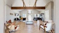 Located at 1125 Old Graves Mill Rd in Lynchburg, Virginia, this single-floor community space offers a generously sized, open-concept layout designed for both gathering and productivity. The centerpiece of the space is a dramatic vaulted ceiling accented by striking exposed wood beams, creating a warm yet contemporary architectural aesthetic throughout the main area. Light wood-tone flooring runs seamlessly underfoot, complementing the neutral gray walls and bright, airy atmosphere achieved through abundant natural light from multiple large windows and sliding glass doors.

The main open area flows effortlessly between a well-appointed lounge zone — featuring comfortable seating arrangements around a wall-mounted television — and a fully equipped kitchen. The kitchen showcases dark cabinetry, stainless steel appliances, a herringbone tile backsplash, granite countertops, and a large island with bar seating, ideal for casual dining or entertaining. A secondary kitchenette area with high-top bistro tables provides an additional casual breakroom setting.

A dedicated conference room offers a streamlined, professional environment with a large wooden table, leather seating, a wall-mounted display, and a whiteboard. Sliding glass doors throughout open to outdoor areas, further extending the sense of space and connection to the surrounding neighborhood.