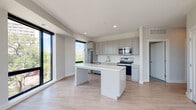 This single-floor residence showcases a clean, contemporary design characterized by a neutral palette, light wood-style flooring, and an abundance of natural light. The open-concept living and kitchen area serves as the heart of the unit, where large black-framed floor-to-ceiling windows frame sweeping views of the surrounding neighborhood, flooding the space with daylight. The kitchen is well-appointed with flat-front gray cabinetry extending to the ceiling, a white quartz countertop, and a full suite of stainless steel appliances including a bottom-freezer refrigerator, dishwasher, microwave, and electric range. A central island with a matching white quartz surface provides additional workspace and casual seating. The living area is generously proportioned, anchored by dramatic corner floor-to-ceiling windows that create a bright, airy atmosphere with open city views. Two bathrooms are finished in the same modern aesthetic, each featuring white quartz vanity tops, undermount sinks with brushed nickel fixtures, large mirrors, recessed lighting, and separate shower areas. The consistent use of light tones and minimalist design elements throughout creates a cohesive, sophisticated living environment.