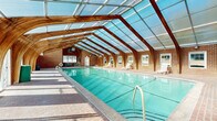 Located at 1401 Eland Downe in Phoenixville, PA, this single-floor indoor pool facility is a standout example of thoughtful architectural design blending function with visual appeal. The centerpiece of the space is a generously sized lap pool with clearly marked lanes, its vivid turquoise water reflecting the abundant natural light that pours in from above.

The most distinctive architectural feature is the sweeping vaulted roof structure, composed of elegant wooden arched beams supporting a translucent polycarbonate panel ceiling. This design floods the entire pool area with diffused daylight, creating a bright and airy atmosphere year-round while maintaining the comfort of an enclosed environment.

The perimeter walls are constructed of classic red brick, lending warmth and durability to the space. Multiple windows and glass doors line the long sides of the building, further enhancing natural light and providing views of the surrounding greenery. White lounge chairs are positioned along the pool deck, offering comfortable spots for relaxation.

A dedicated seating and rest area near the entrance features a brick-paved floor, arched brick detailing, and a garden bench, providing a welcoming transitional space. Safety equipment, including a life ring, is conveniently mounted on the wall. The overall ambiance is bright, open, and inviting, making this facility well-suited for both recreational swimming and structured aquatic activities.