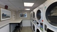 This communal laundry facility is designed with practicality and comfort in mind, offering residents a clean and organized space for their laundry needs. The room is lined with multiple stacked washer and dryer units along one wall, providing ample capacity to accommodate a high volume of laundry. The machines are modern and well-maintained, ensuring efficient and reliable performance.

The space is well-lit by large recessed panel lights set into a drop ceiling, creating a bright and welcoming atmosphere. A dedicated folding table with chairs is conveniently positioned in the room, giving users a comfortable surface to sort and fold laundry. A framed artwork piece on the wall adds a touch of character to the otherwise utilitarian space.

The walls feature a clean, neutral palette with a tiled wainscoting detail, while the light-toned tile flooring contributes to the overall sense of cleanliness. A payment kiosk is also present, suggesting a convenient and cashless or card-based system for operating the machines. The single-floor layout ensures easy accessibility, making this laundry facility a straightforward and efficient amenity for all users.