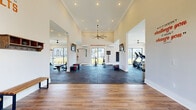 This single-floor fitness center is thoughtfully designed to support a full range of workout styles within a bright, open layout. The space is organized into distinct zones, each tailored to specific training needs. The strength training area is outfitted with a power rack, weight plates, adjustable benches, dumbbells, kettlebells, and medicine balls, all set on durable rubber flooring. A dedicated cardio section features treadmills, stationary bikes, and rowing machines, positioned alongside floor-to-ceiling windows that overlook a sparkling pool and outdoor lounge area, flooding the space with natural light. A functional training zone offers TRX suspension systems, plyo boxes, and resistance equipment for versatile workouts. Throughout the space, bold motivational phrases such as "Hustle for Muscle," "Harder. Better. Faster. Stronger." and "Big Results" are displayed on the crisp white walls, reinforcing an energetic atmosphere. Ceiling fans maintain airflow across all zones, complementing the climate control systems. Large mirrors amplify the sense of space, while wall-mounted televisions provide entertainment. The combination of soaring ceilings, abundant natural light, and premium equipment makes this fitness center a standout amenity.