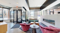 Located in Saint Paul, Minnesota, the Mods Lobby, Gathering, and Lounge is a thoughtfully designed communal space that seamlessly blends contemporary architecture with eclectic interior styling. The space is organized into several distinct yet connected zones, each offering its own character and function.

The main lobby entrance features polished concrete floors, a glass-enclosed vestibule entry, and floor-to-ceiling windows that frame views of the surrounding streetscape, flooding the interior with natural light. A reception desk anchors the entry area, while nearby seating—including plush crimson swivel chairs and a tufted green sofa—creates a welcoming lounge vibe.

Moving deeper into the space, a gathering lounge showcases soaring double-height ceilings with striking vertical slat ceiling details in dark wood. A linear fireplace set into a concrete wall adds warmth, while bold furnishings, including a teal velvet sofa and gold-accented coffee table, lend a curated, upscale feel. A large wall-mounted television makes this area ideal for both relaxation and socializing.

A café-style area features warm brick walls, a white quartz island with bar seating, and a dark mirrored backsplash. High-top tables with red upholstered stools provide casual seating beneath a sculptural pendant light. A secondary corridor lounge features patterned wallcovering, wingback chairs, and polished tile flooring, offering a quieter retreat within the broader space.