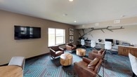 This single-floor amenity space is a thoughtfully designed communal area that seamlessly blends lounge, dining, and coworking functions into one open, flowing layout. The interior is anchored by a striking teal and brown geometric-patterned carpet that runs throughout, lending a bold, contemporary identity to the space.

The lounge area is furnished with rich brown leather sofas and armchairs arranged around natural wood stump accent tables, creating a warm and inviting gathering environment. Elegant multi-arm chandeliers with cylindrical shades hang overhead, complementing the recessed lighting embedded in the smooth white ceilings. Large windows along the perimeter flood the space with natural light and offer open views of the surrounding landscape.

Multiple round dining tables with tufted and upholstered chairs are positioned throughout, offering versatile settings for casual meals or small group meetings. A dedicated business center lines one wall, featuring a long white desk with computer workstations set beneath a sculptural branch-style wall installation, ideal for residents or guests seeking a productive workspace.

Glass-panel doors and dark-framed architectural details add a modern edge, while warm taupe wall tones and curated abstract artwork maintain a polished, cohesive aesthetic throughout. The space is designed to accommodate a variety of social and professional activities in an open, welcoming environment.