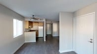 This studio presents a practical and well-organized open-concept layout that makes efficient use of its space. The flooring throughout is a dark hardwood-style material that adds a modern contrast to the light gray walls and white trim, creating a clean and cohesive aesthetic.

The kitchen is positioned along one wall and features warm honey-toned wooden cabinetry, a white refrigerator, and a sink with a dark countertop. A peninsula-style counter extends outward, providing additional prep space and a casual dining or workspace area. A ceiling fan with a light fixture is installed above the kitchen area, offering both ventilation and ambient lighting.

The textured popcorn ceiling runs throughout the space, adding a classic touch, while neutral gray walls keep the interior feeling open and versatile. A window near the kitchen allows natural light to filter into the living area.

A hallway visible from the main living area leads toward additional spaces, including what appears to be a closet and a separate room, providing a sense of depth and functional separation within the studio's layout. Overall, the space offers a straightforward and functional living environment.