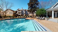 Located in Daphne, Alabama, this outdoor pool area offers a well-appointed retreat designed for relaxation and leisure. The centerpiece is a generously sized swimming pool with crystal-clear blue water, accessible via wide entry steps fitted with handrails. The surrounding pool deck is finished in warm-toned pavers, providing ample space for sunbathing and socializing, lined with multiple lounge chairs and shaded seating areas complete with patio umbrellas.

Mature magnolia trees and lush, manicured landscaping frame the space, offering natural shade and a sense of privacy. A decorative wrought-iron fence borders the perimeter, defining the area while maintaining an open, airy feel under the bright Alabama sky.

Adjacent to the pool, a covered porch features classic white columns and stacked stone accents at the base, lending a refined, Southern architectural character. A row of French doors with multi-pane windows lines the porch wall, flooding the interior with views of the pool area. Charming rocking chairs and ceiling fans complete the porch, creating a comfortable shaded respite. The overall ambiance is inviting and resort-like, blending classic Southern design elements with practical outdoor amenities suited for warm-weather enjoyment.