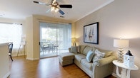 Located at 1600 Belle Point Dr in the desirable community of Mt. Pleasant, South Carolina, this single-floor residence offers a comfortable and well-appointed living environment. The living area is a standout feature, showcasing warm hardwood flooring that adds a sense of warmth and continuity throughout the space. Crown molding lines the ceiling, lending a polished, refined touch to the room's aesthetic. A ceiling fan with integrated lighting anchors the space overhead, providing both comfort and functionality. Natural light pours in through a full-height sliding glass door, which opens directly onto a private balcony furnished with outdoor seating — an ideal spot to enjoy the lush, tree-lined surroundings. Vertical blinds offer flexible light control and privacy. The living space is generously proportioned, easily accommodating a sectional sofa and additional seating. Neutral, earthy wall tones create a calm and inviting atmosphere that complements the natural finishes throughout. The overall ambiance is bright, airy, and relaxed — characteristic of the coastal Lowcountry lifestyle that Mt. Pleasant is known for. This residence blends practical comfort with tasteful design details in a well-situated South Carolina location.