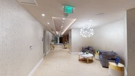 This single-floor wellness spa is designed with a cohesive, upscale aesthetic that prioritizes comfort and relaxation throughout. The space is laid out along an elongated corridor with herringbone-patterned light wood flooring that runs continuously, unifying the various zones and lending warmth to the overall environment.

The reception and lounge area serves as the welcoming centerpiece, featuring a sculptural starburst chandelier as a focal point above a seating arrangement of plush gray velvet chairs and a round marble-top coffee table with gold-toned legs. Recessed lighting and a coffered ceiling detail with subtle ambient uplighting add dimension and a polished finish to this area.

Moving deeper into the space, a striking living green wall introduces a bold natural element, providing a vibrant contrast against the otherwise neutral tones. Adjacent to it, a full-body massage chair station offers an immediate relaxation option. A slatted wood partition elegantly divides areas while maintaining an open, airy feel, and a branded floor mat marks the transition into the inner wellness zones.

The steam room is enclosed by full-height glass panels with brass-finished hardware, offering a sleek, contemporary look. Inside, tiered mosaic-tiled benches line the walls, creating a functional and visually refined steam experience. Recessed lighting illuminates the space cleanly. The overall ambiance throughout is calm, polished, and purposefully designed for a premium wellness experience.