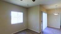 This single-story space presents a clean, functional layout with a welcoming ambiance defined by its warm beige walls and wood-style hard flooring. The room pictured is generously proportioned, offering an open feel well-suited for a variety of uses. Natural light is a defining feature, with two large windows fitted with vertical blinds — one positioned along the side wall and another framing what appears to be a secondary area or passageway — allowing diffused light to brighten the interior throughout the day. A ceiling fan is centrally installed, providing practical climate comfort. The walls are finished in a smooth, neutral tone that complements the warm flooring beneath, creating a cohesive and understated aesthetic. White baseboards provide a clean contrast against both the walls and flooring. Electrical outlets are conveniently placed throughout. The overall condition of the space appears well-maintained, with a straightforward, move-in ready character. The light, airy atmosphere is enhanced by the abundance of natural light filtering through the window treatments, giving the room an open and inviting quality.