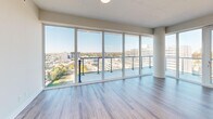 This modern high-rise unit offers a thoughtfully designed layout across a single floor, comprising three bedrooms, two bathrooms, a combined kitchen and living area, and a dedicated closet. Light hardwood flooring flows consistently throughout the main living spaces, complementing the clean, contemporary aesthetic defined by crisp white walls and abundant natural light.

The open-concept kitchen is equipped with a full suite of stainless steel appliances, including a refrigerator, range, microwave, and dishwasher. White cabinetry extends to the ceiling for generous storage, while a central island provides additional workspace. A distinctive sculptural pendant light fixture serves as a focal point above the island. The living area is anchored by an expansive wall of floor-to-ceiling windows with balcony access, offering sweeping panoramic city and treetop views.

Each bedroom features the same light hardwood flooring and large windows that frame elevated urban views. The primary bedroom includes access to a dedicated walk-in closet. The two bathrooms are finished with light marble-style tile flooring and white vanities, with one bathroom housing a glass-enclosed walk-in shower alongside an in-unit stacked washer and dryer. The overall design prioritizes clean lines, neutral tones, and maximized natural light throughout.