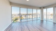 This modern high-rise unit offers a thoughtfully designed layout across a single floor, comprising three bedrooms, two bathrooms, a combined kitchen and living area, and a dedicated closet. Light hardwood flooring flows consistently throughout the main living spaces, complementing the clean, contemporary aesthetic defined by crisp white walls and abundant natural light.

The open-concept kitchen is equipped with a full suite of stainless steel appliances, including a refrigerator, range, microwave, and dishwasher. White cabinetry extends to the ceiling for generous storage, while a central island provides additional workspace. A distinctive sculptural pendant light fixture serves as a focal point above the island. The living area is anchored by an expansive wall of floor-to-ceiling windows with balcony access, offering sweeping panoramic city and treetop views.

Each bedroom features the same light hardwood flooring and large windows that frame elevated urban views. The primary bedroom includes access to a dedicated walk-in closet. The two bathrooms are finished with light marble-style tile flooring and white vanities, with one bathroom housing a glass-enclosed walk-in shower alongside an in-unit stacked washer and dryer. The overall design prioritizes clean lines, neutral tones, and maximized natural light throughout.