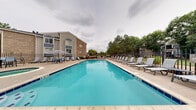 Located at 1724 Robb St in Lakewood, Colorado, this outdoor pool area offers a well-appointed recreational retreat designed for relaxation and leisure. The centerpiece is a generously sized rectangular swimming pool with vivid turquoise water, framed by a wide concrete sundeck lined with rows of cushioned lounge chairs — ideal for sunbathing and unwinding. A separate circular hot tub provides an additional relaxation option, positioned prominently on the deck near umbrella-shaded seating arrangements with tables and chairs for outdoor dining or socializing. The pool area is fully enclosed by a black metal fence, ensuring privacy and security, while mature trees along the perimeter provide natural shade and a pleasant green backdrop. A dedicated pool house structure on the premises houses mechanical equipment and utility access, with posted pool rules and notices clearly visible. Overhead lighting is provided for evening use. The surrounding landscape features lush, tall trees that enhance the outdoor ambiance and offer a sense of seclusion. The multi-story residential buildings visible beyond the fence complement the community-oriented character of the space. Overall, the pool area is thoughtfully designed to accommodate a range of outdoor activities in a clean, organized, and inviting environment in the heart of Lakewood, CO.