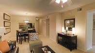 This single-floor residence offers an open-concept layout that integrates the living, dining, and kitchen areas into one cohesive, flowing space. The living area is anchored by a ceiling fan and features warm beige walls and light-toned carpeting, creating a comfortable and inviting atmosphere. A dark-finished console table with shelving adds a stylish storage element, complemented by a large mirror that reflects light and enhances the sense of space.

The dining area is defined by a dark wood table and matching chairs, illuminated by a classic pendant light that adds a warm, focused glow. Adjacent to the dining space, the kitchen features dark cabinetry and appliances visible in the background, providing a functional contrast to the lighter tones of the surrounding areas.

A central hallway leads toward what appears to be a closet and additional rooms, maintaining a practical flow throughout the unit. White doors and trim throughout offer clean visual definition against the warm wall tones. The consistent flooring and unified color palette across all visible areas contribute to a harmonious, well-coordinated interior that feels both functional and welcoming.