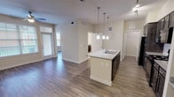 This single-story, one-bedroom, one-bathroom residence offers a thoughtfully designed open-concept floor plan finished with warm, neutral tones and wood-style flooring that flows throughout the main living areas. The kitchen serves as a central feature of the space, equipped with dark espresso-toned shaker-style cabinetry extending to the upper walls, granite-style countertops, and a full suite of black appliances including a gas range, microwave, and refrigerator. A peninsula island with a built-in sink and pendant lighting overhead provides additional prep space and casual seating, connecting seamlessly to the open living area. The living room is generously sized, featuring a ceiling fan, track lighting, and built-in shelving, creating a functional yet comfortable environment. Large windows in both the living area and bedroom invite natural light and outdoor views. The bedroom is well-proportioned with its own ceiling fan and ample natural light. The bathroom includes a full soaking tub with tiled surround and light tile flooring. A dedicated laundry area with a full-size washer and dryer and built-in shelving adds everyday convenience to the overall layout.